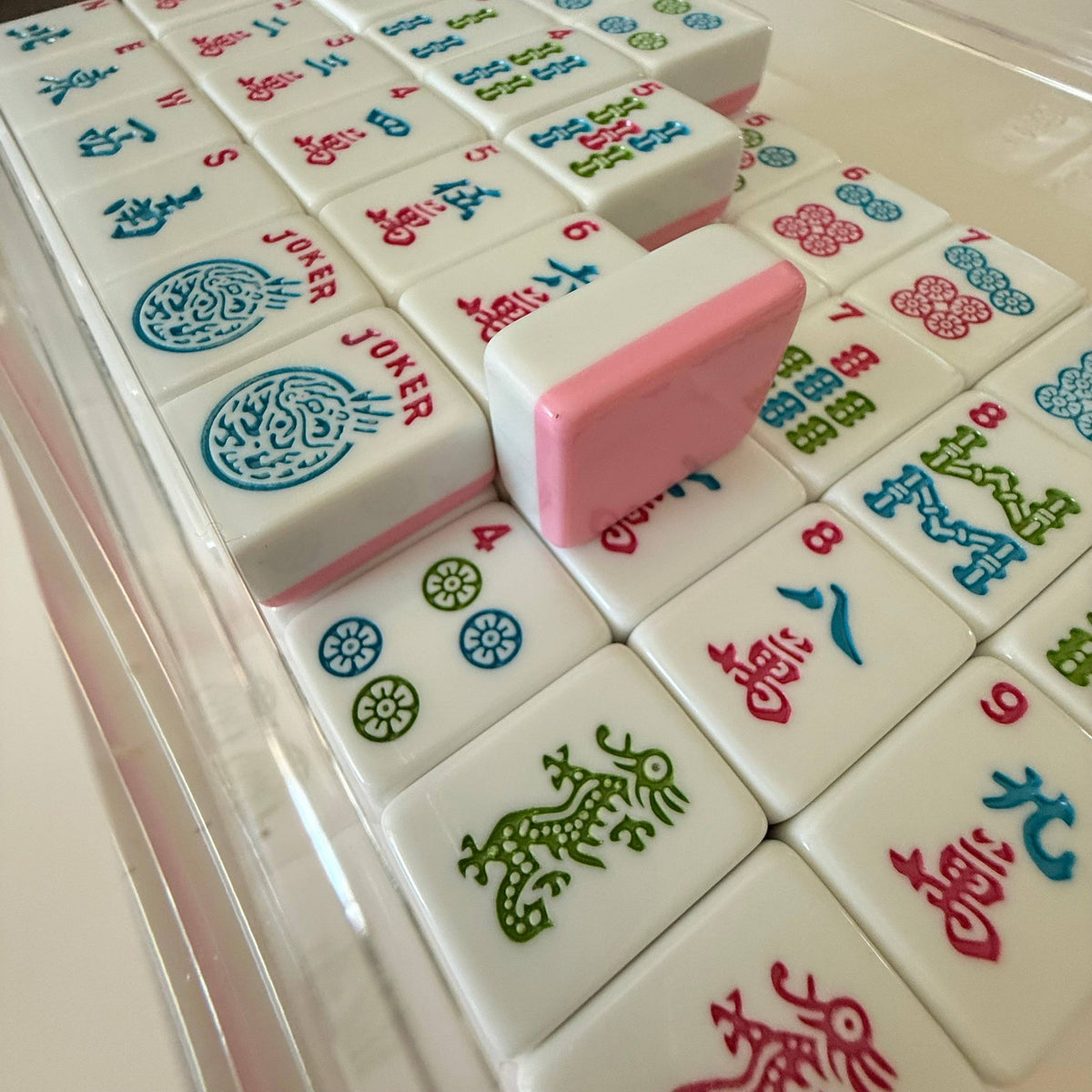 A close-up of a Mahjong set inside a clear plastic storage case, showing multiple white tiles with colorful Chinese characters and a pink-and-white tile resting on top.